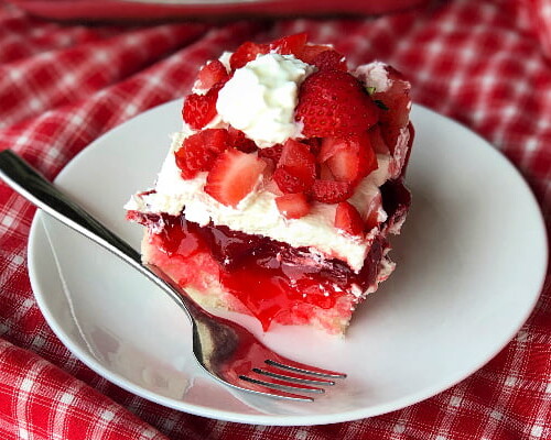 A piece of layered strawberry poke cake with cool whip on a white plate with a fork on the side.