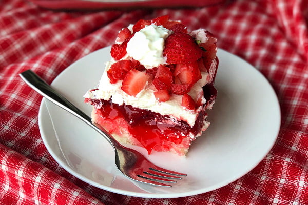 A piece of layered strawberry poke cake with cool whip on a white plate with a fork on the side.