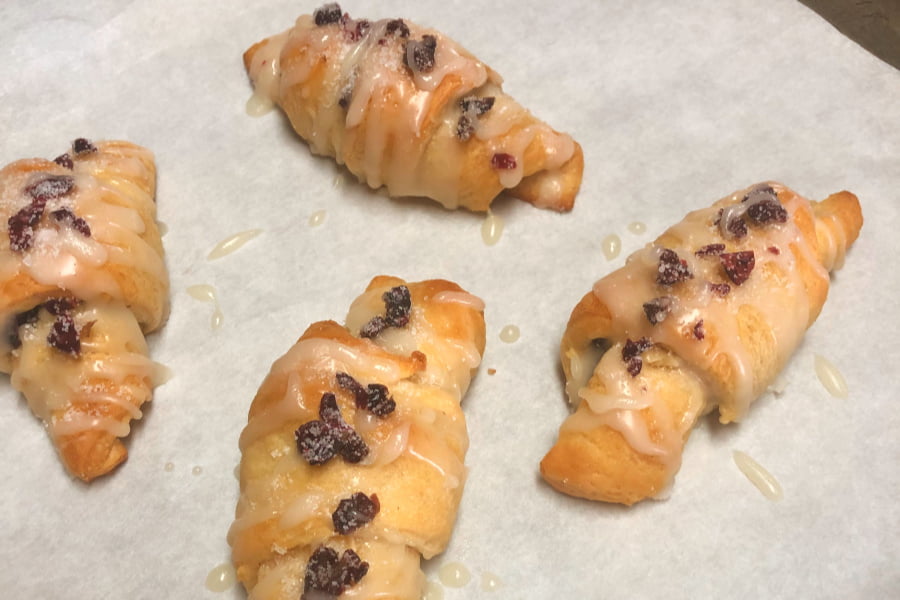 Baking sheet of baked crescent rolls that are filled with dried cranberries and chopped walnuts, then drizzled with a powdered sugar glaze.