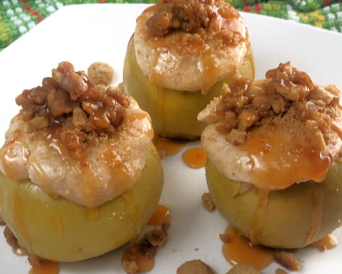 Three cheesecake-stuffed baked apples, drizzled with caramel sauce, sitting on a white plate.