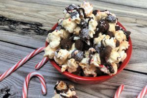 Red bowl of kettle corn munch with chocolate cookies, dark chocolate drizzle and crushed peppermint. Candy canes are shown around bowl of munch.