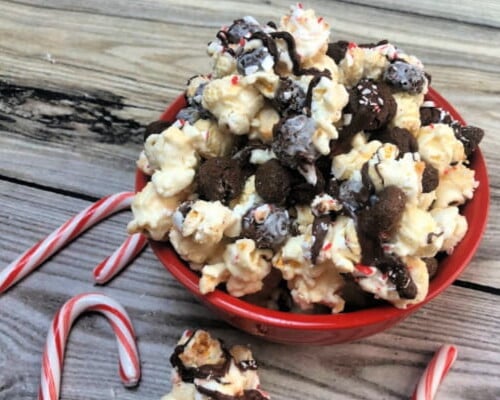 Red bowl of kettle corn munch with chocolate cookies, dark chocolate drizzle and crushed peppermint. Candy canes are shown around bowl of munch.