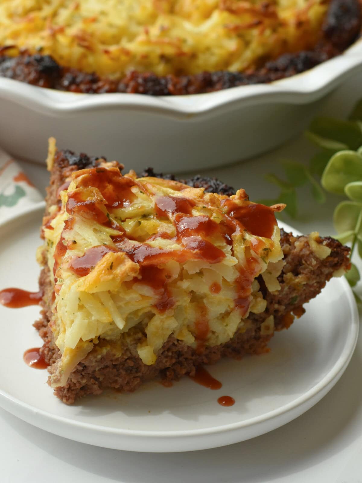 meatloaf-pie-bbq-sauce A slice of meatloaf pie with cheesy hashbrowns and a a drizzle of BBQ sauce.