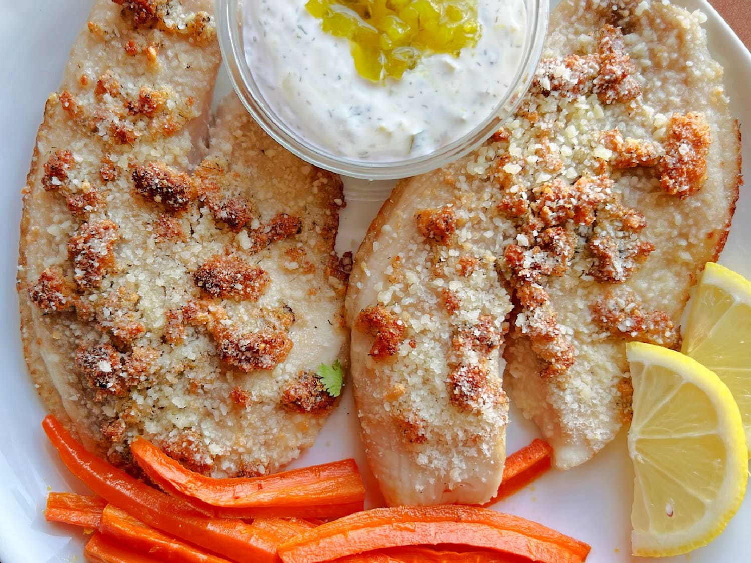 Two air fryer tilapia fillets with tartar sauce and lemons on the side. 