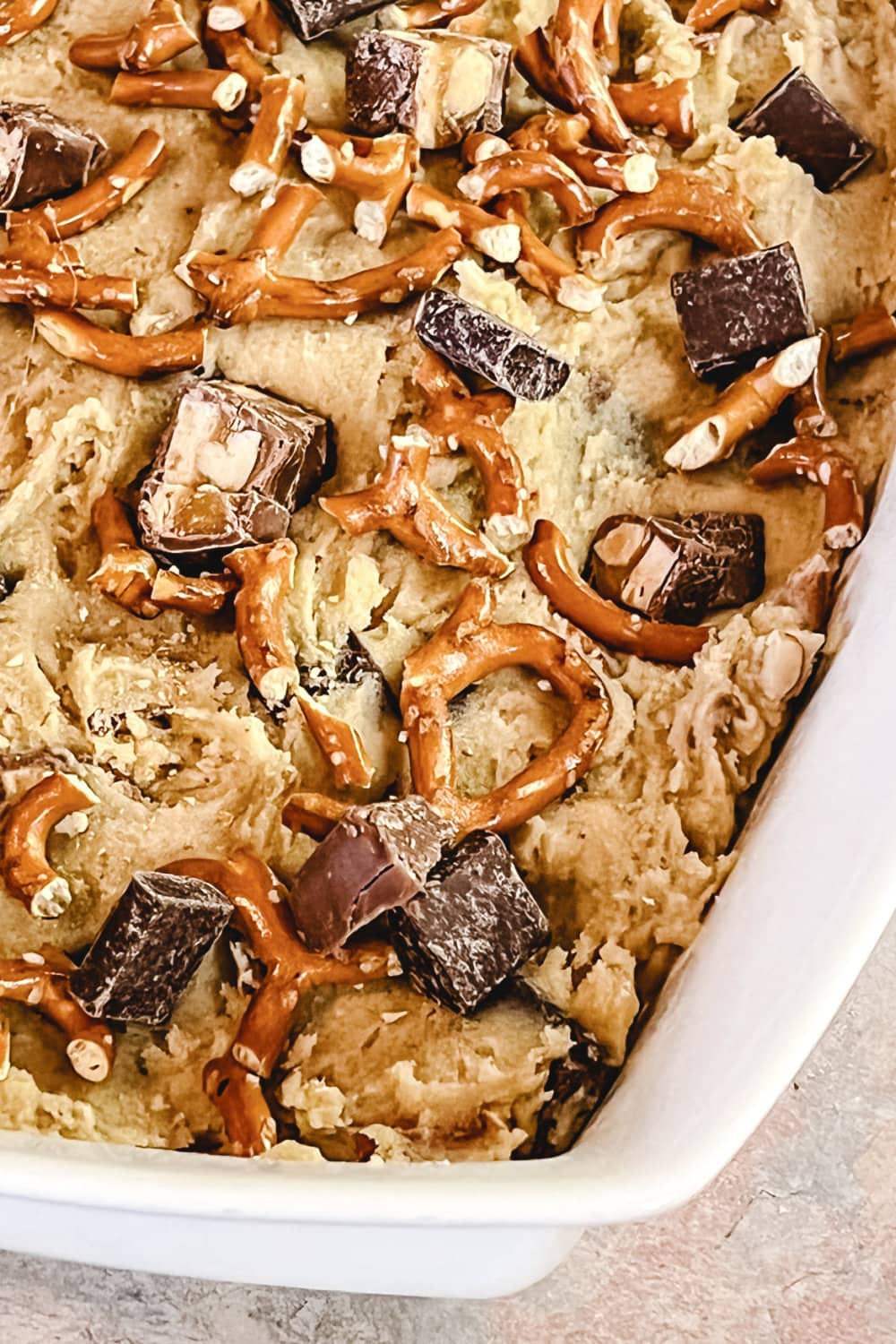 Cookie bar dough in a baking pan, topped with chocolate chunks and pretzels. 