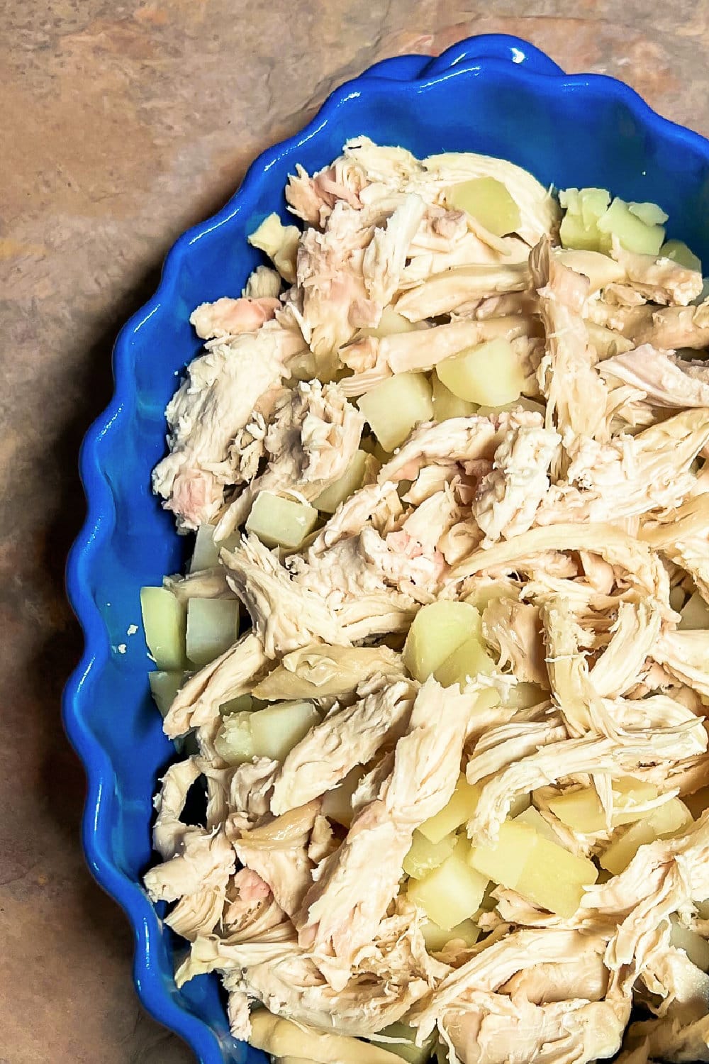 Cooked shredded chicken and diced potatoes in a blue casserole dish. 