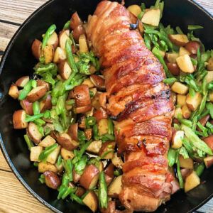 Bacon-wrapped roast pork tenderloin on a bed of potatoes and green beans.