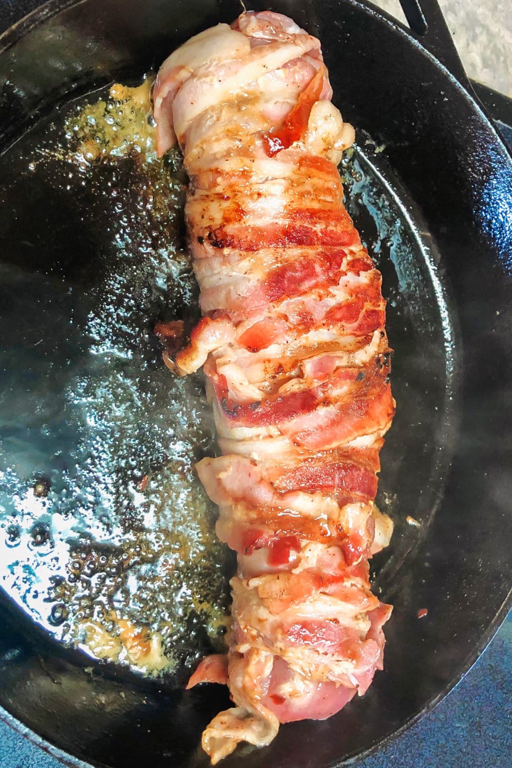 Bacon-wrapped pork tenderloin searing in a skillet. 