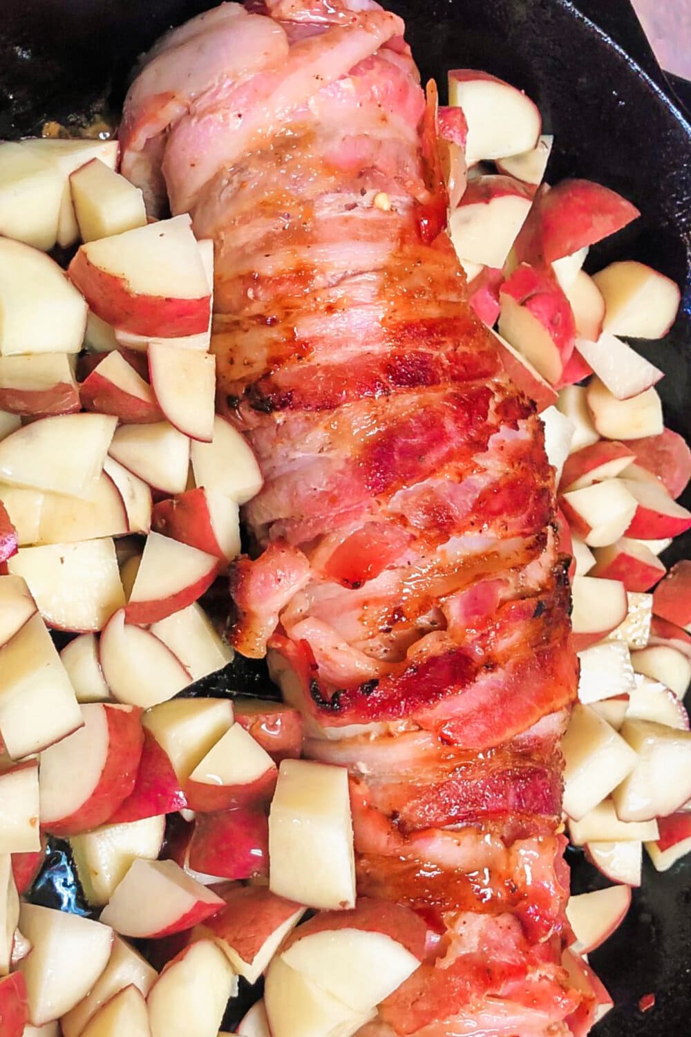 Bacon-wrapped pork tenderloin in a skillet surrounded by raw potatoes. 