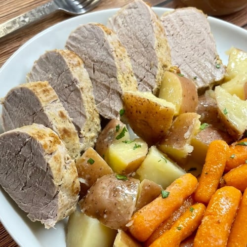 Sliced pork tenderloin on a white place alongside potatoes and baby carrots.