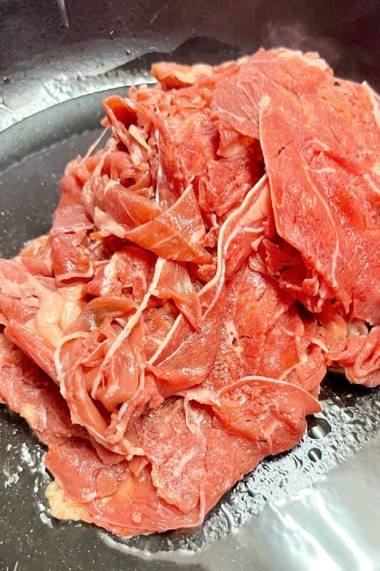 Fresh shaved steak added to hot oil in a skillet to cook. 