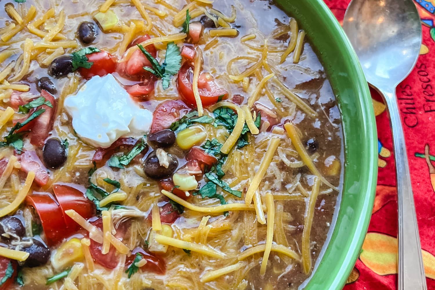 Chicken tortilla soup topped with cheese, tomatoes, and sour cream in a green bowl with a spoon. 