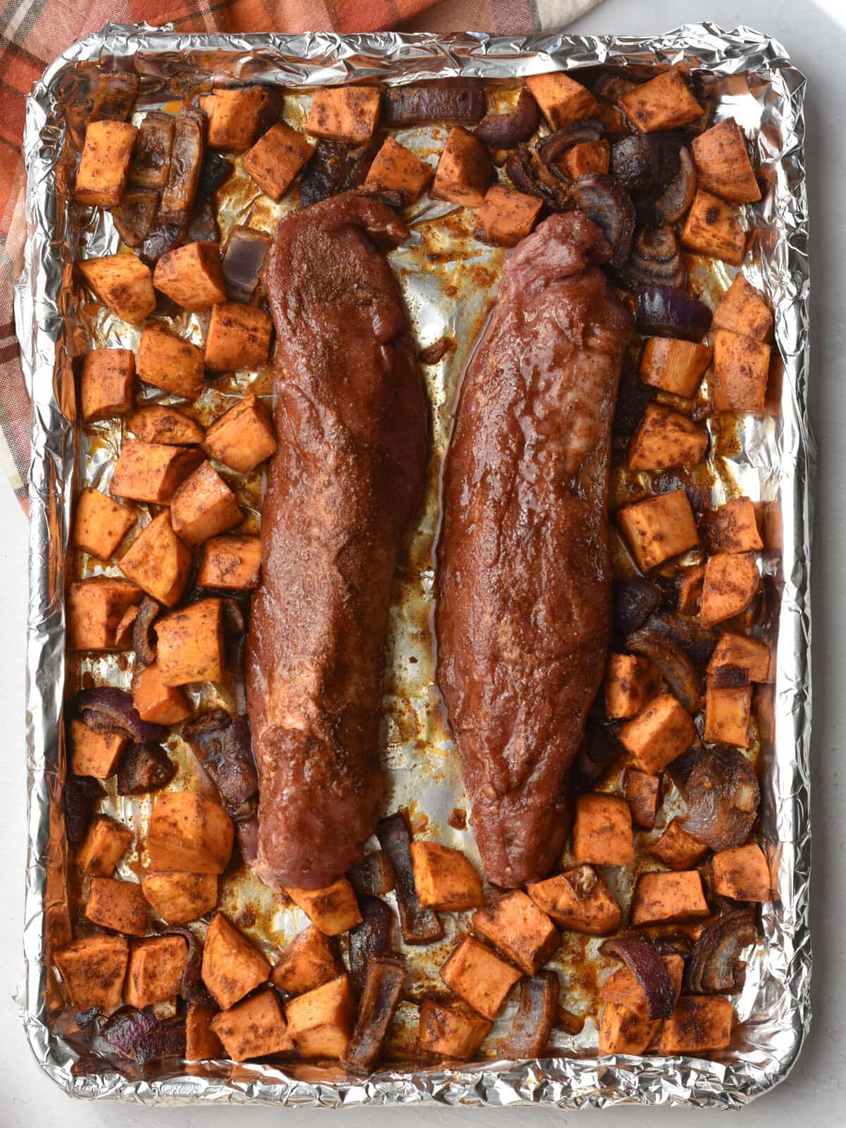 Uncooked pork tenderloin on a sheet pan with seasoned potatoes and red onions. 