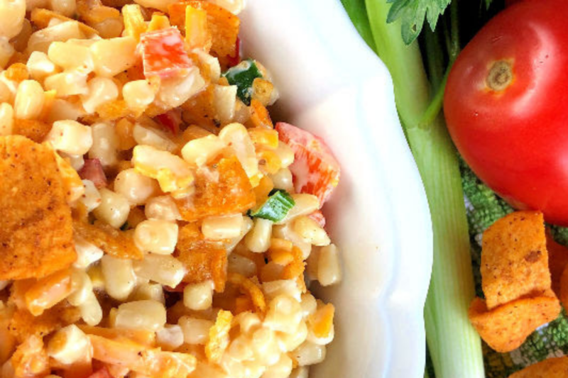 Corn Salad with Chili Cheese Fritos in a white bowl with tomatoes, celery and fritos beside it. 