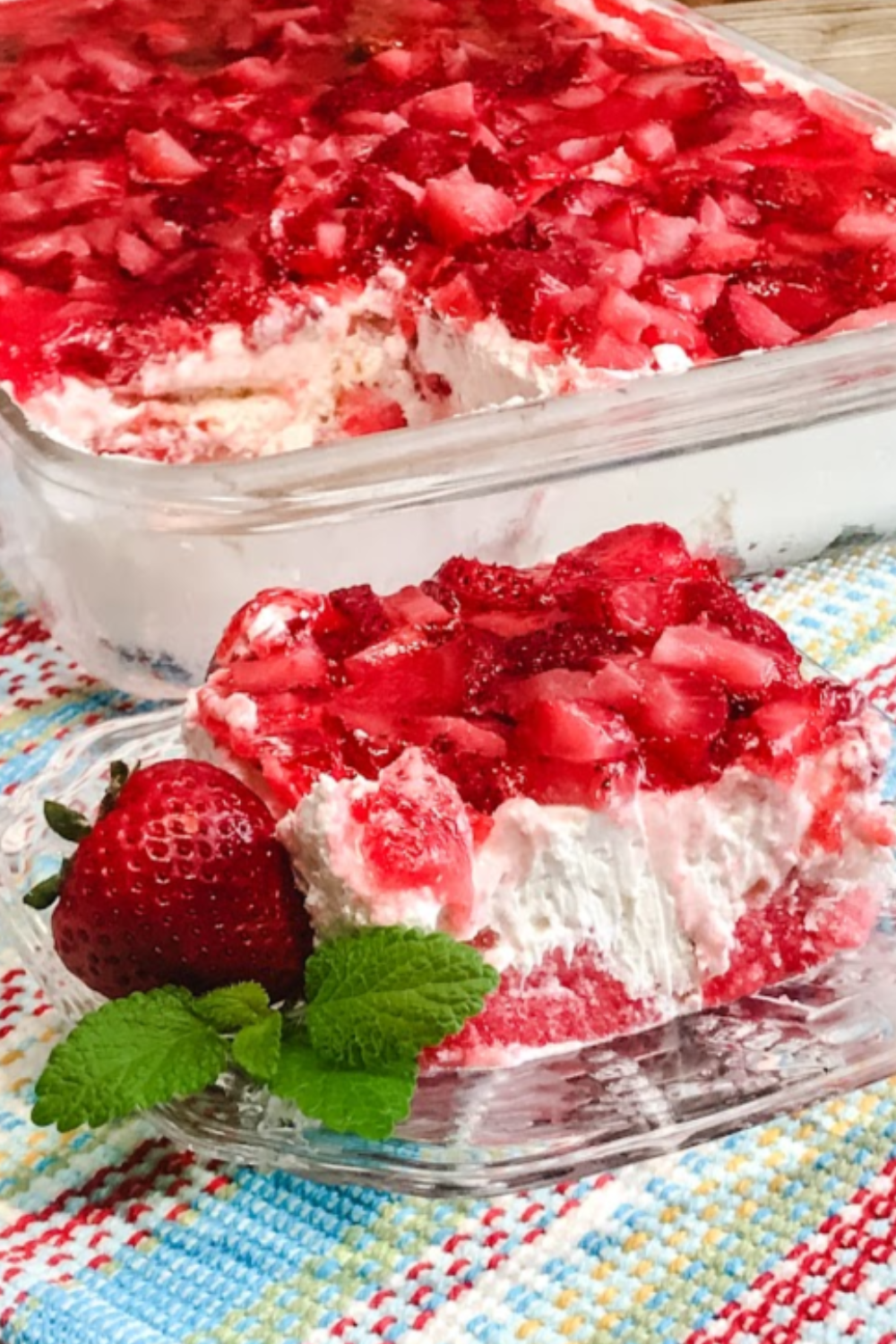 A slice of potluck angel food cake dessert with strawberries sliced on top with the full dish behind it. 