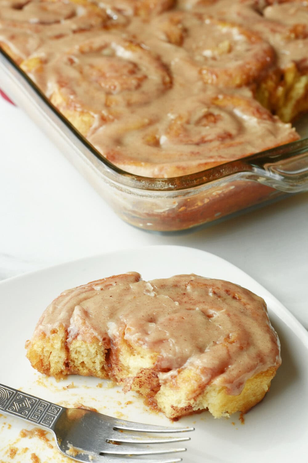 A single pumpkin cinnamon roll with pumpkin spice on a white plate with a fork with a casserole dish behind it. 