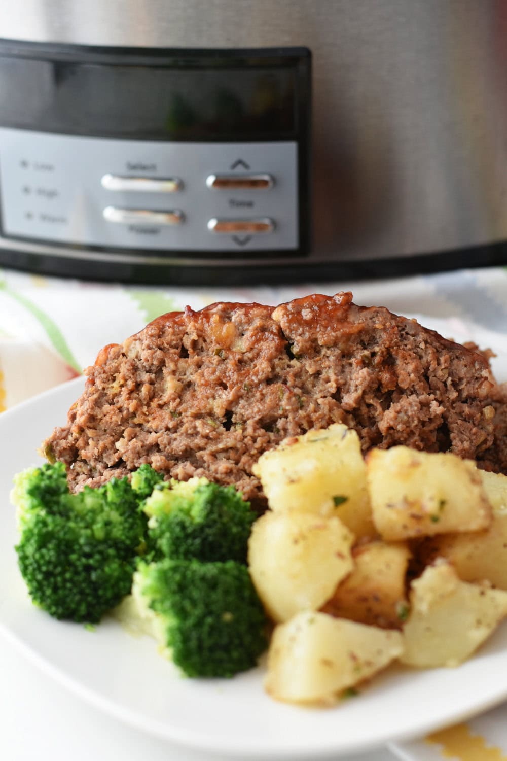 A plate of slow cooker meatloaf with broccoli and potatoes on a white plate with a crock pot behind it. 