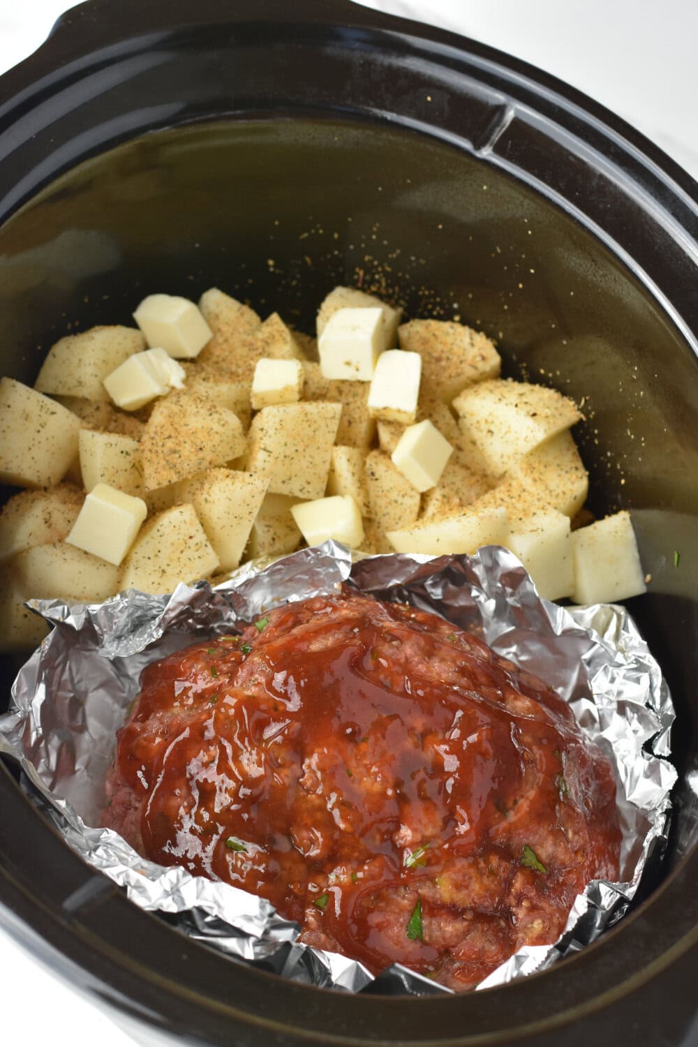 Meatloaf wrapped in foil and cubed potatoes in a slow cooker. 