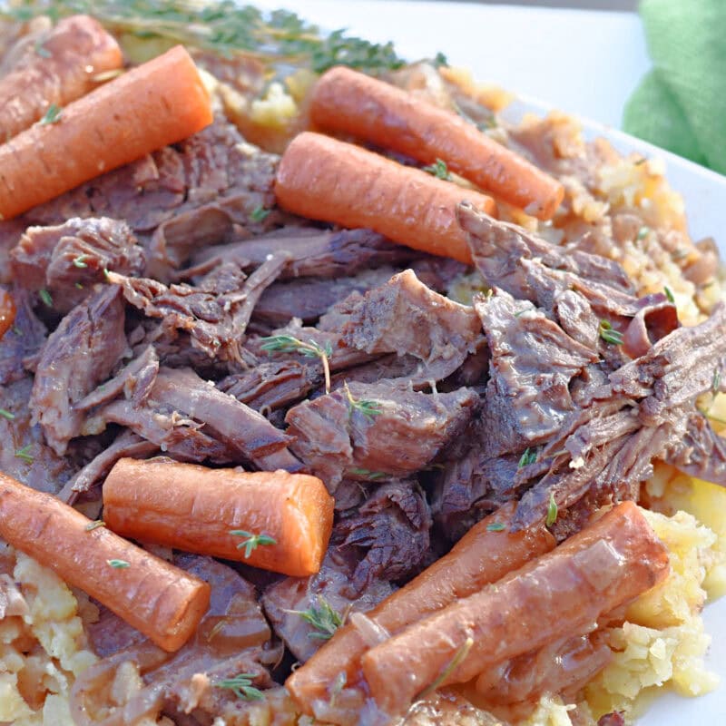 Braised chuck roast with carrots and potatoes on a white plate with a green towel behind it.