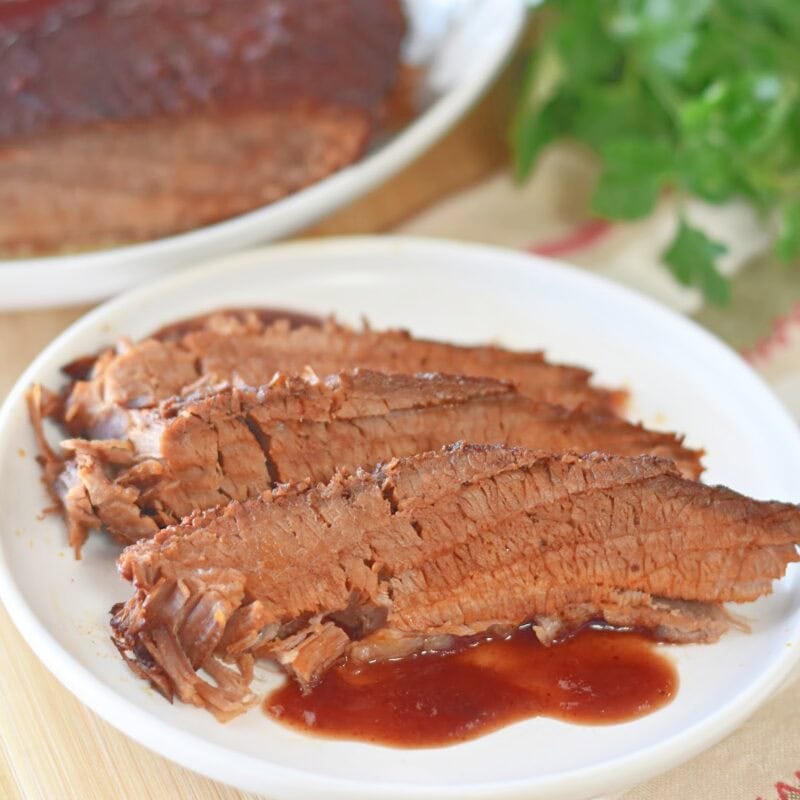 Sliced beef brisket on a white plate with a whole brisket behind it.