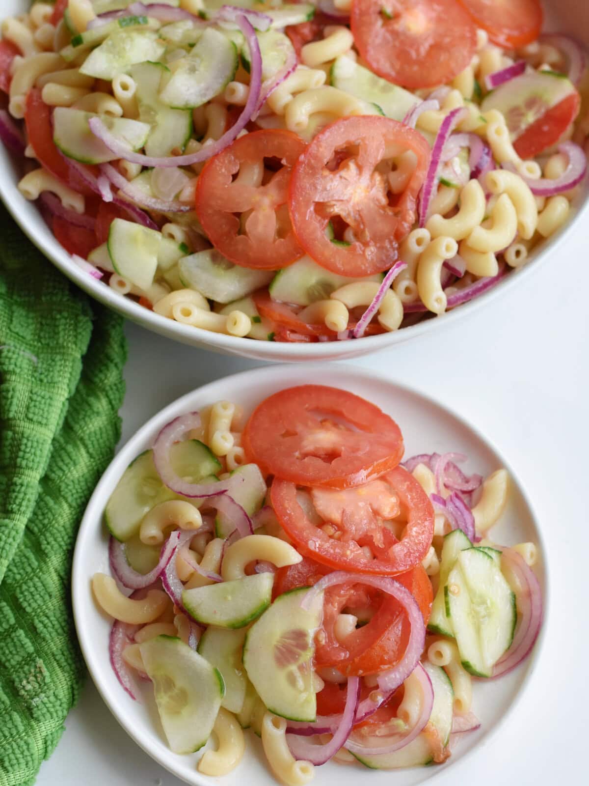 A serving of cucumber tomato pasta salad on a plate with a bowl of the salad behind it. 