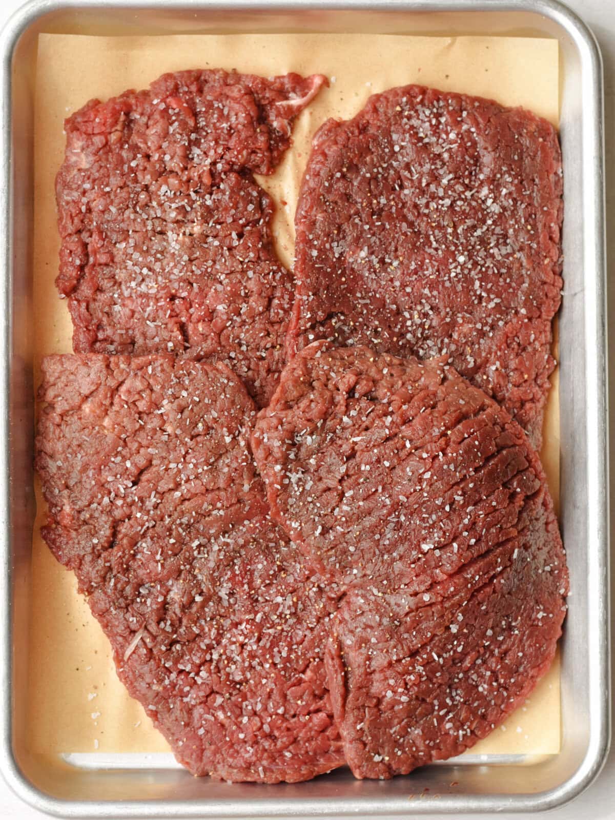 crockpot-round-steak-seasoned Uncooked thin sliced beef that has been tenderized on a baking sheet.
