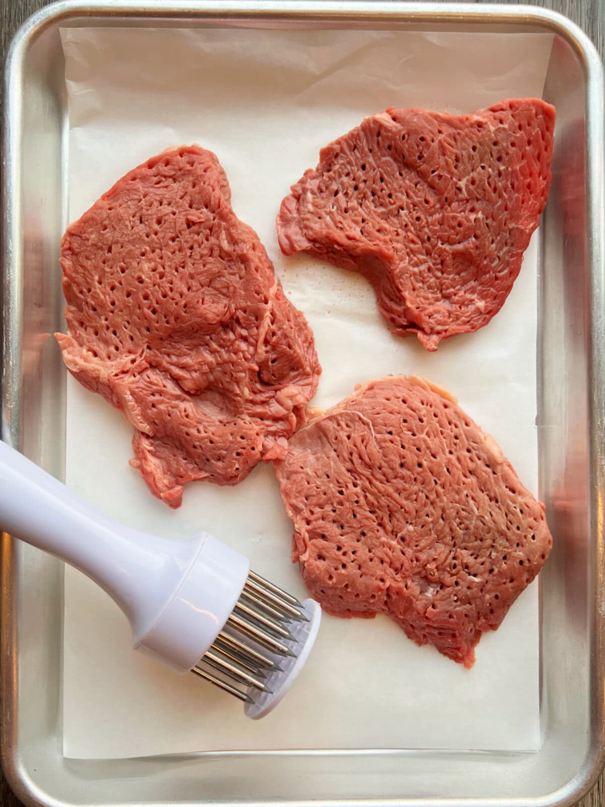 crockpot-round-steak-tenderizer A handheld meat tenderizer with beef steaks in a roasting pan.