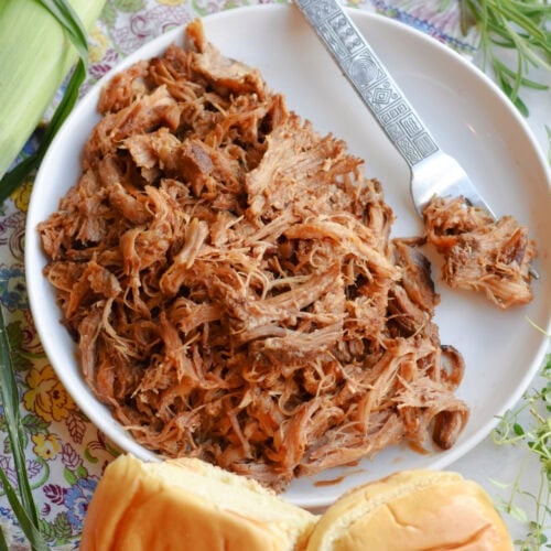 Slow cooker pulled pork on a white plate with white buns beside it.